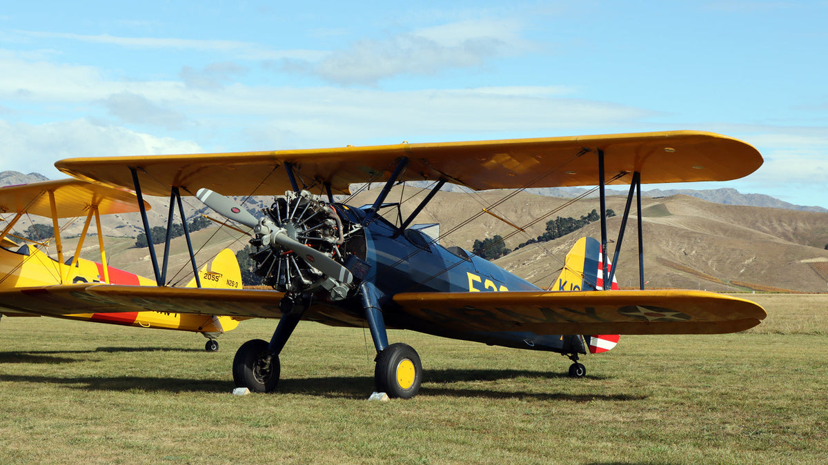 A wide shot of a classic Boeing-Stearman Model 75 ''winnie'' biplane painted in navy blue and bright yellow, parked on a grassy airfield.