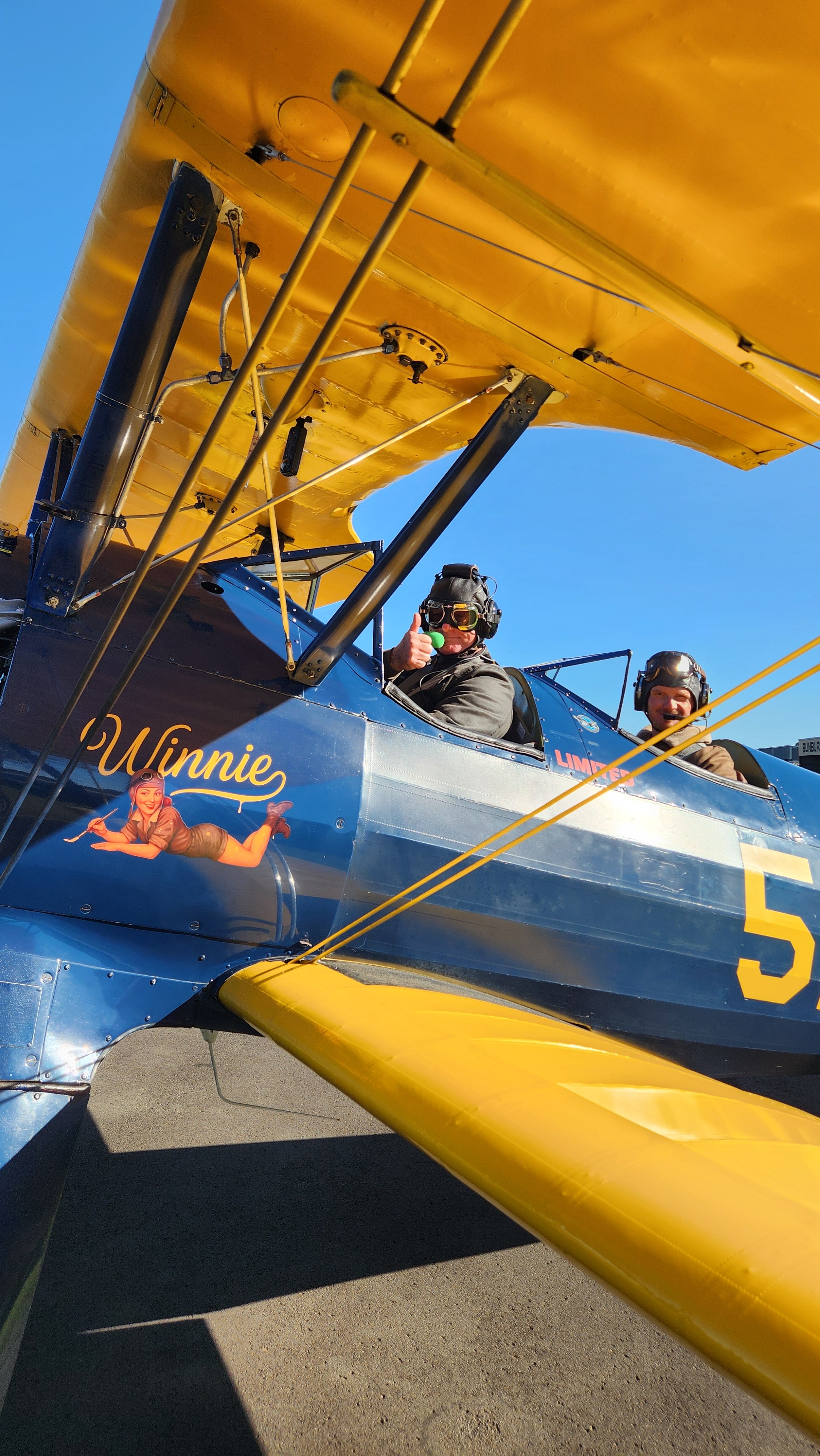 Two people sitting in open cockpits of yellow and blue Boeing-Stearman biplane giving thumbs up.