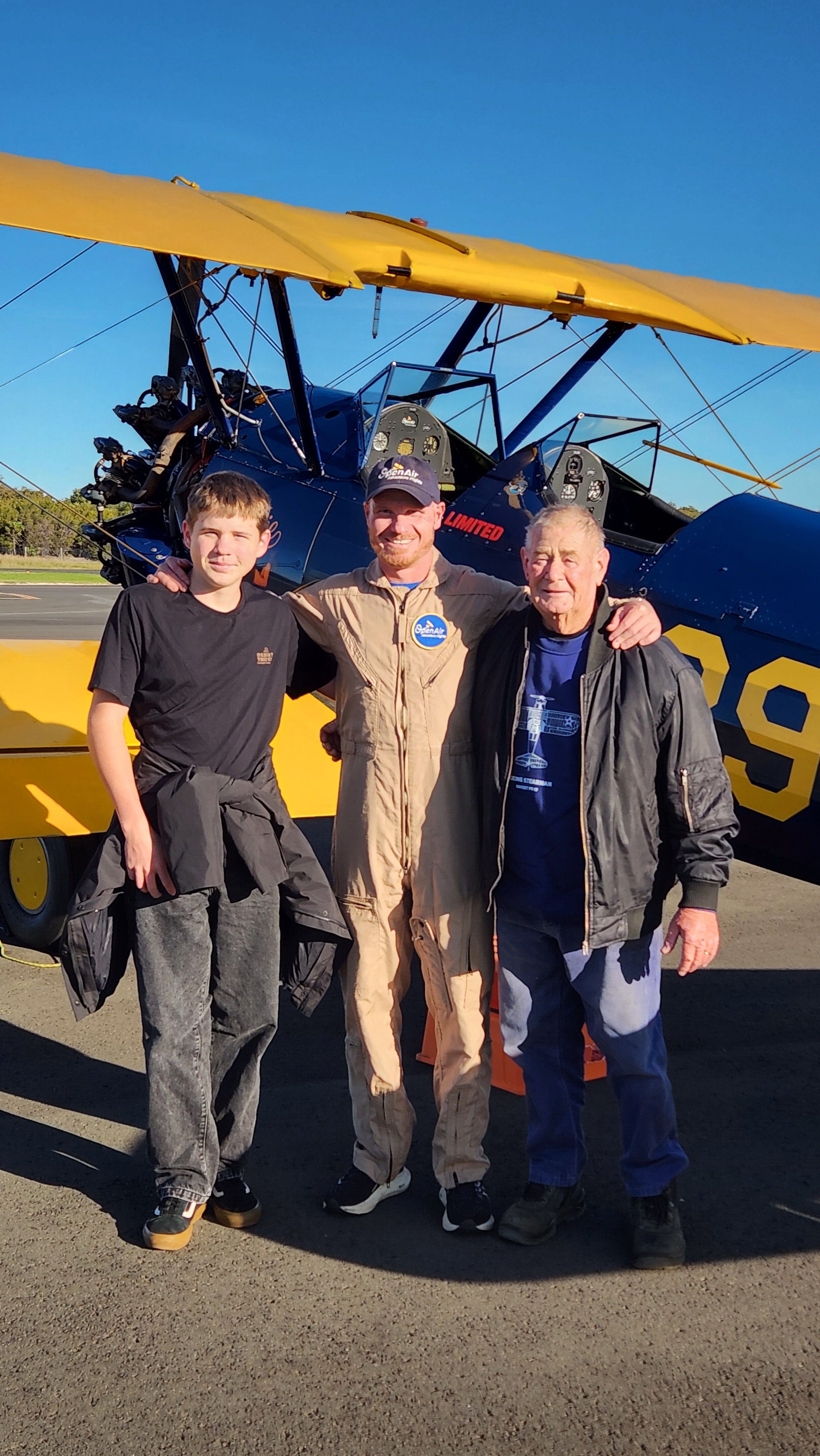 Pilot in flight suit standing with two male passengers on airport tarmac next to yellow and blue Boeing-Stearman biplane.