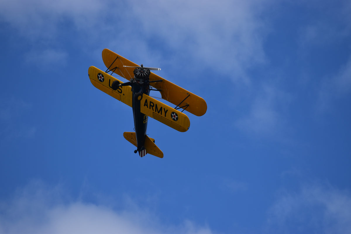 An underside view of a vintage Boeing-Stearman Model 75 biplane banking sharply during flight on a bright blue sky filled with scattered white clouds.