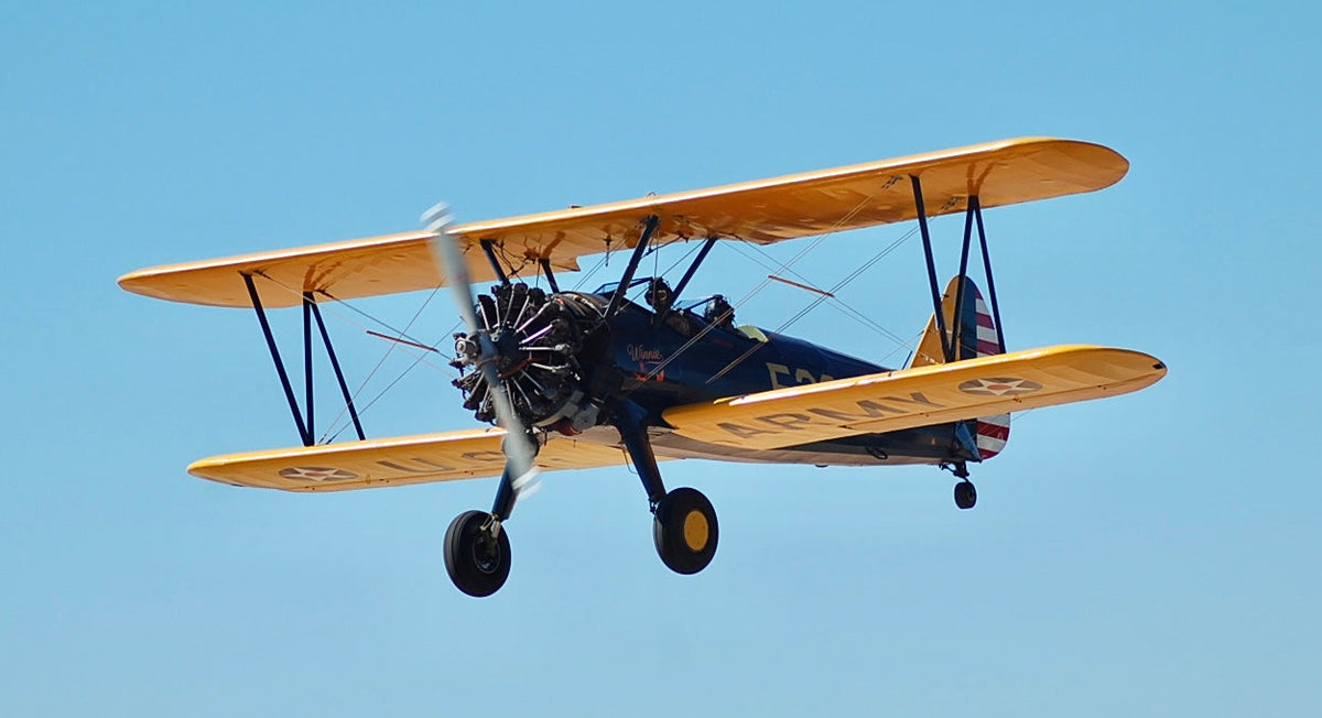 A dynamic front three-quarter view of the vintage Boeing-Stearman Model 75 biplane "Winnie" in mid-flight against a clear blue sky.
