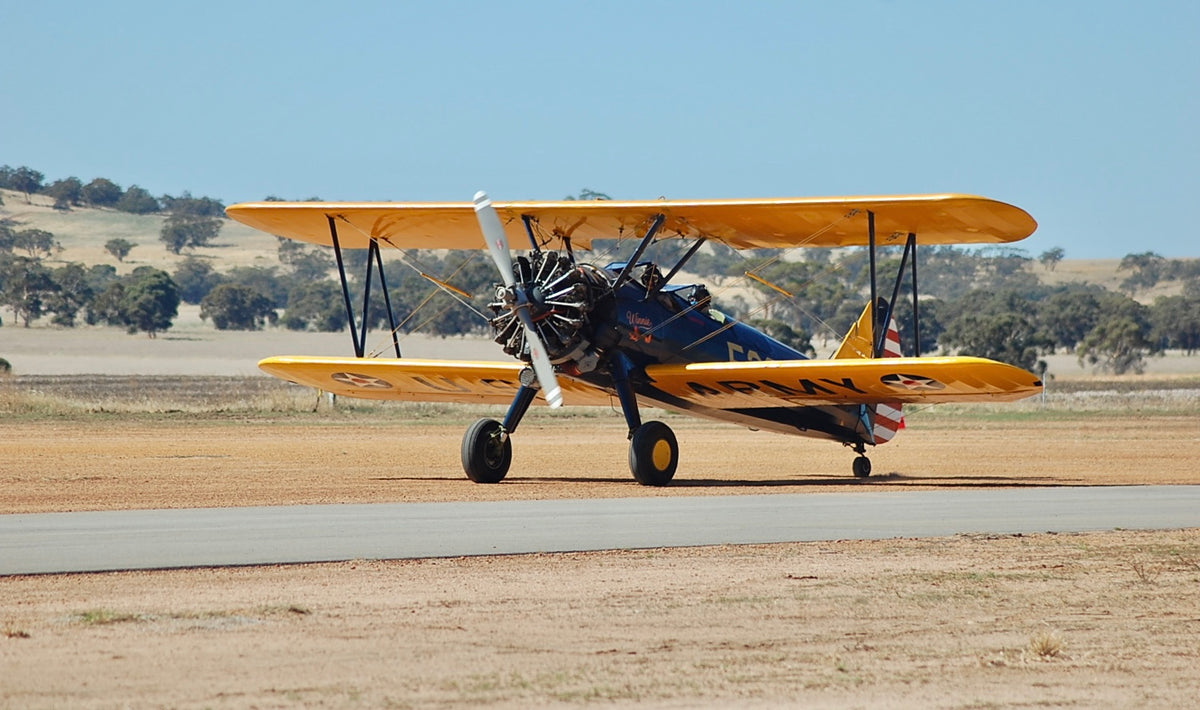 A vintage yellow and blue Boeing-Stearman ''winnie'' biplane idling on an asphalt runway with rolling hills in the background.