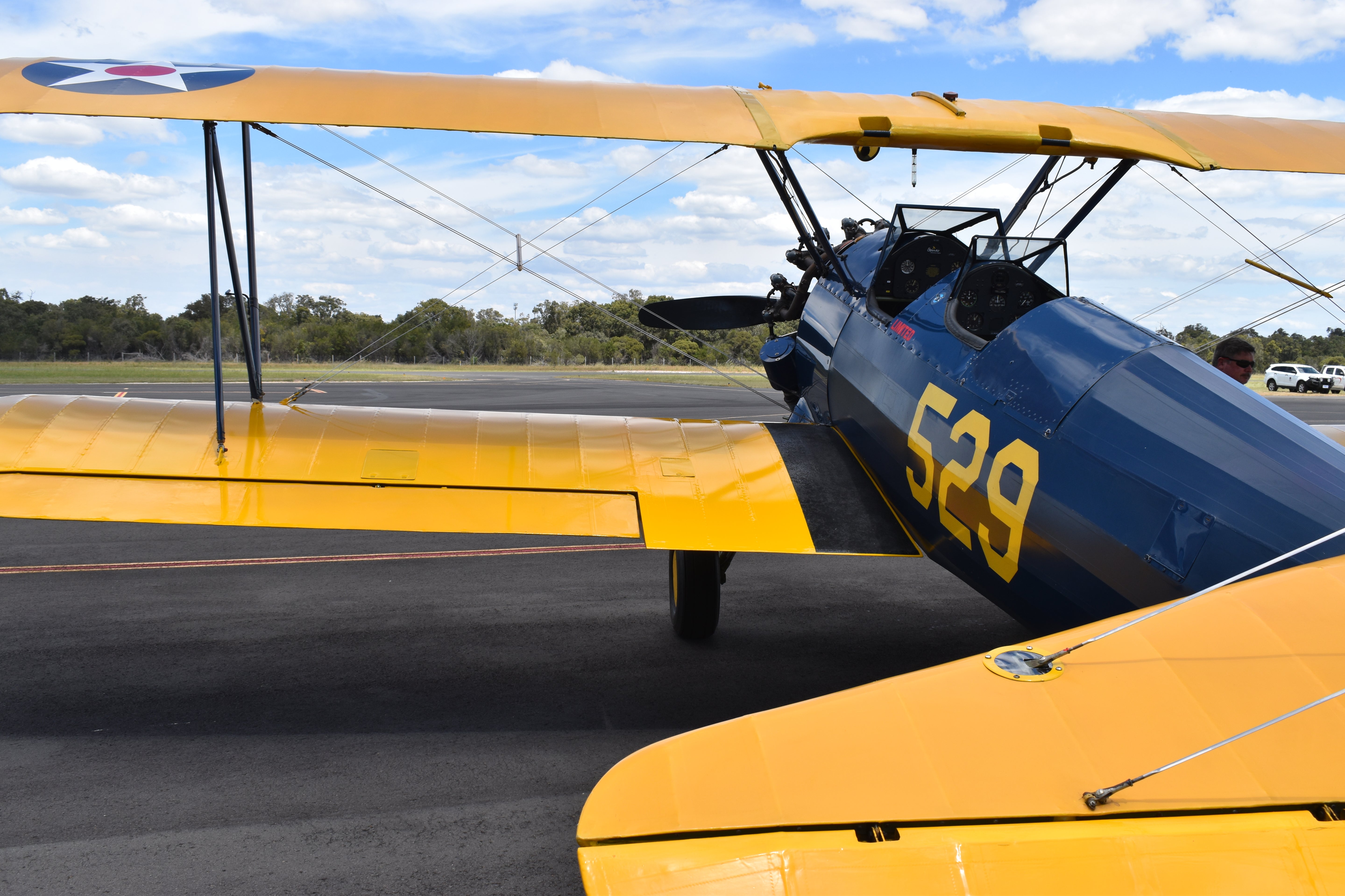 Yellow and blue Boeing Stearman biplane on a runway with trees and sky in the background