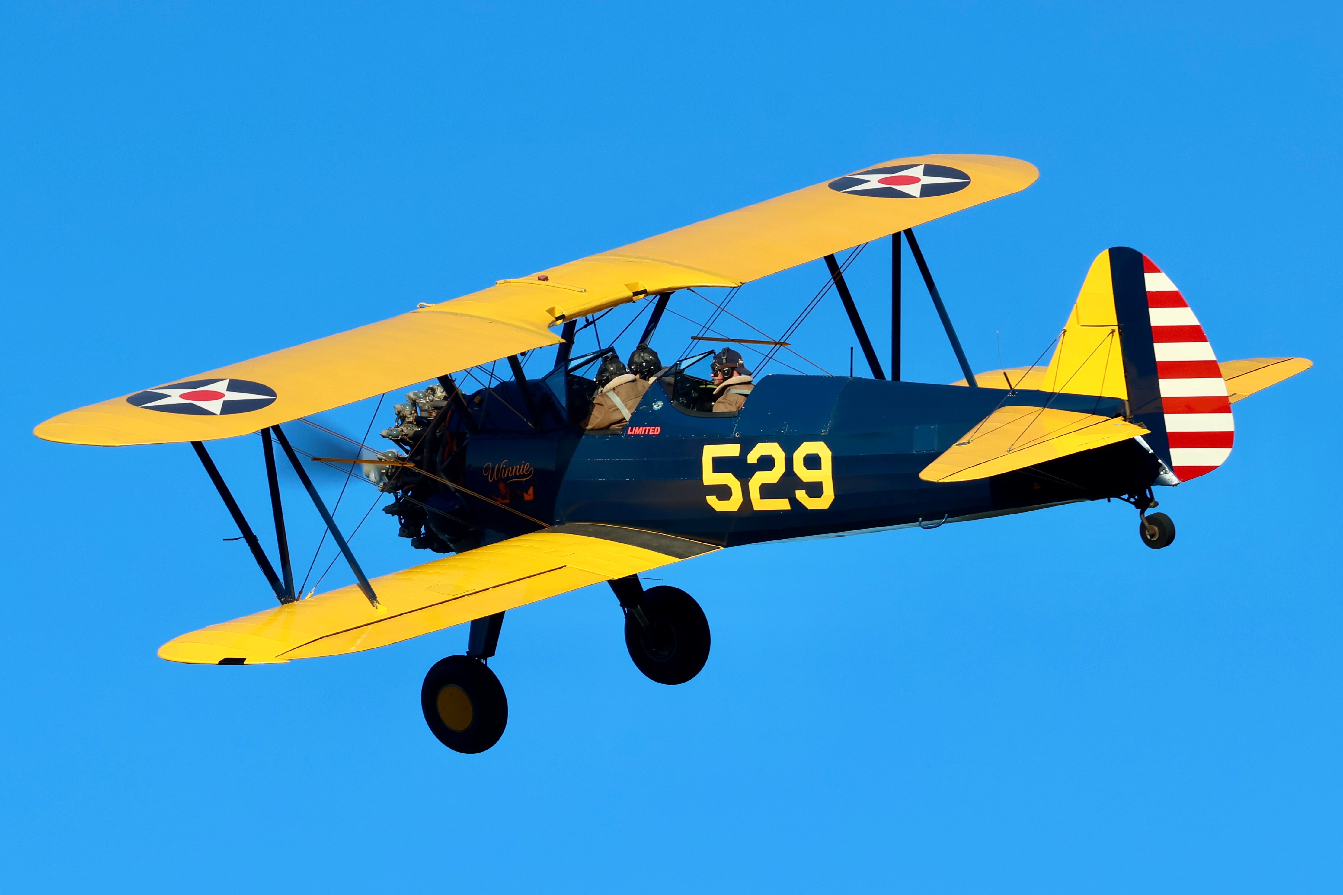 Vintage yellow and blue Boeing-Stearman biplane in flight against clear blue sky.