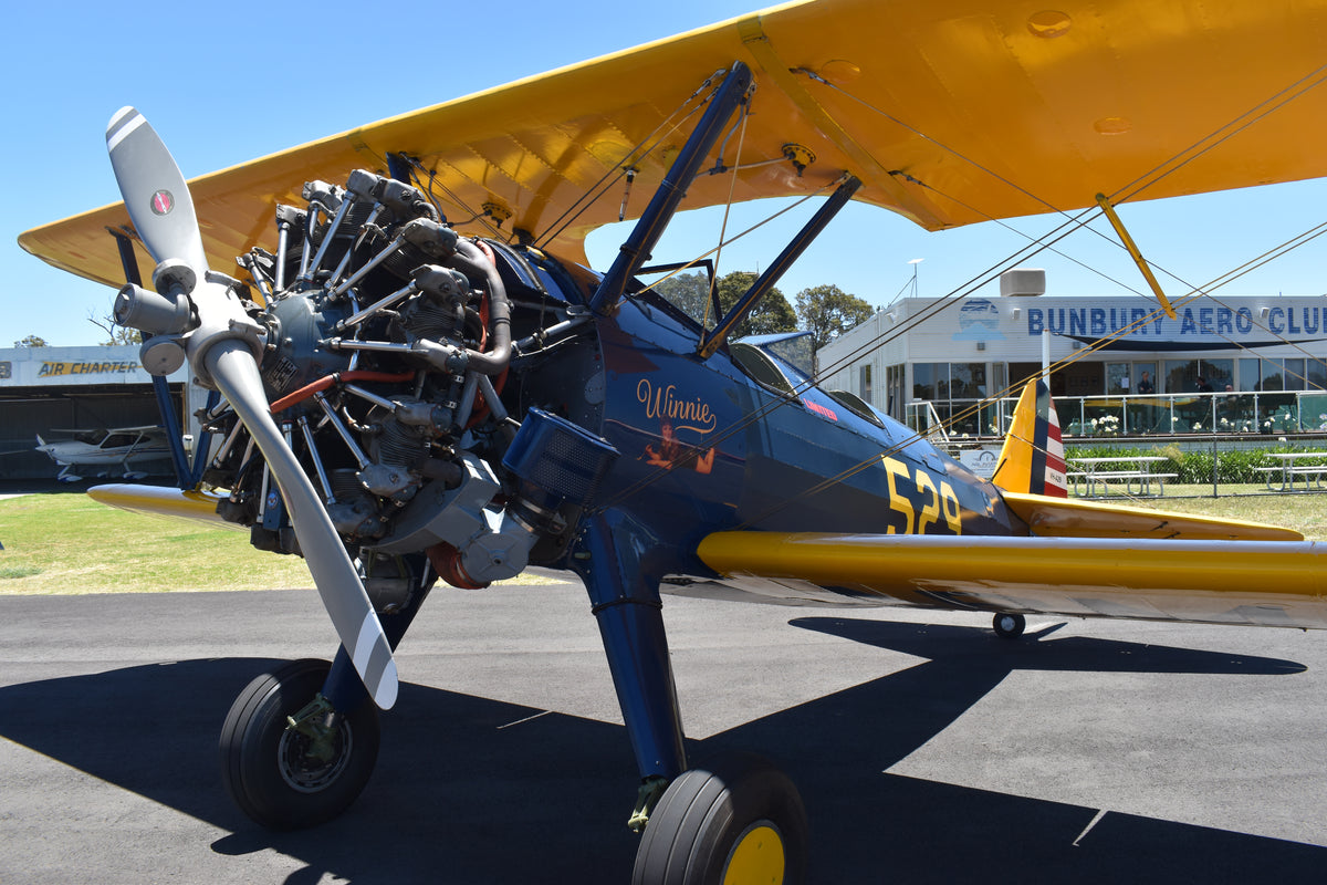 A wide-angle, side-profile view of the classic Boeing-Stearman Model 75 biplane "Winnie" parked on the tarmac outside the Bunbury Aero Club clubhouse. 