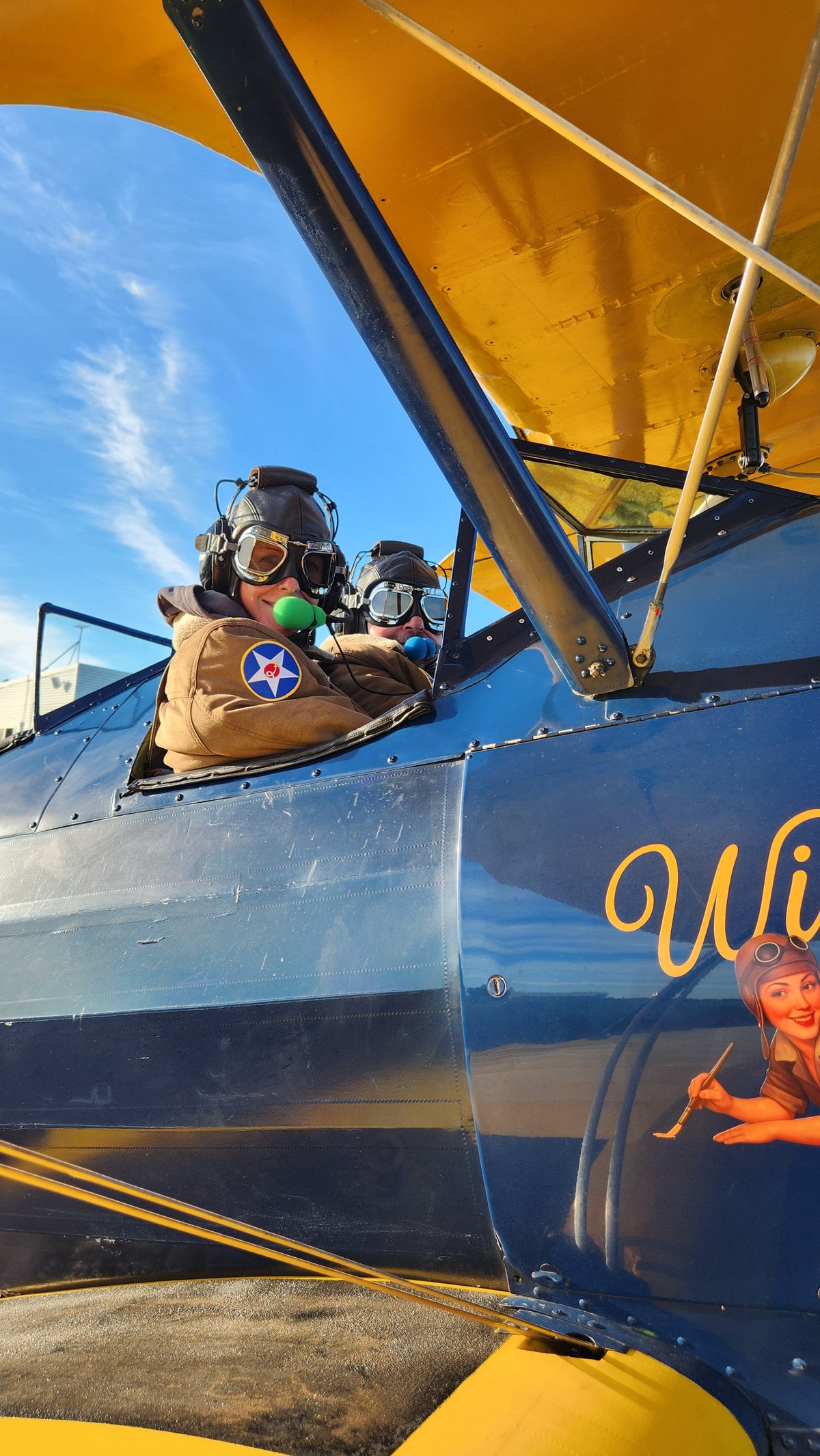 Two people wearing vintage leather flight helmets and goggles sitting inside open cockpits of yellow and blue Boeing-Stearman biplane.