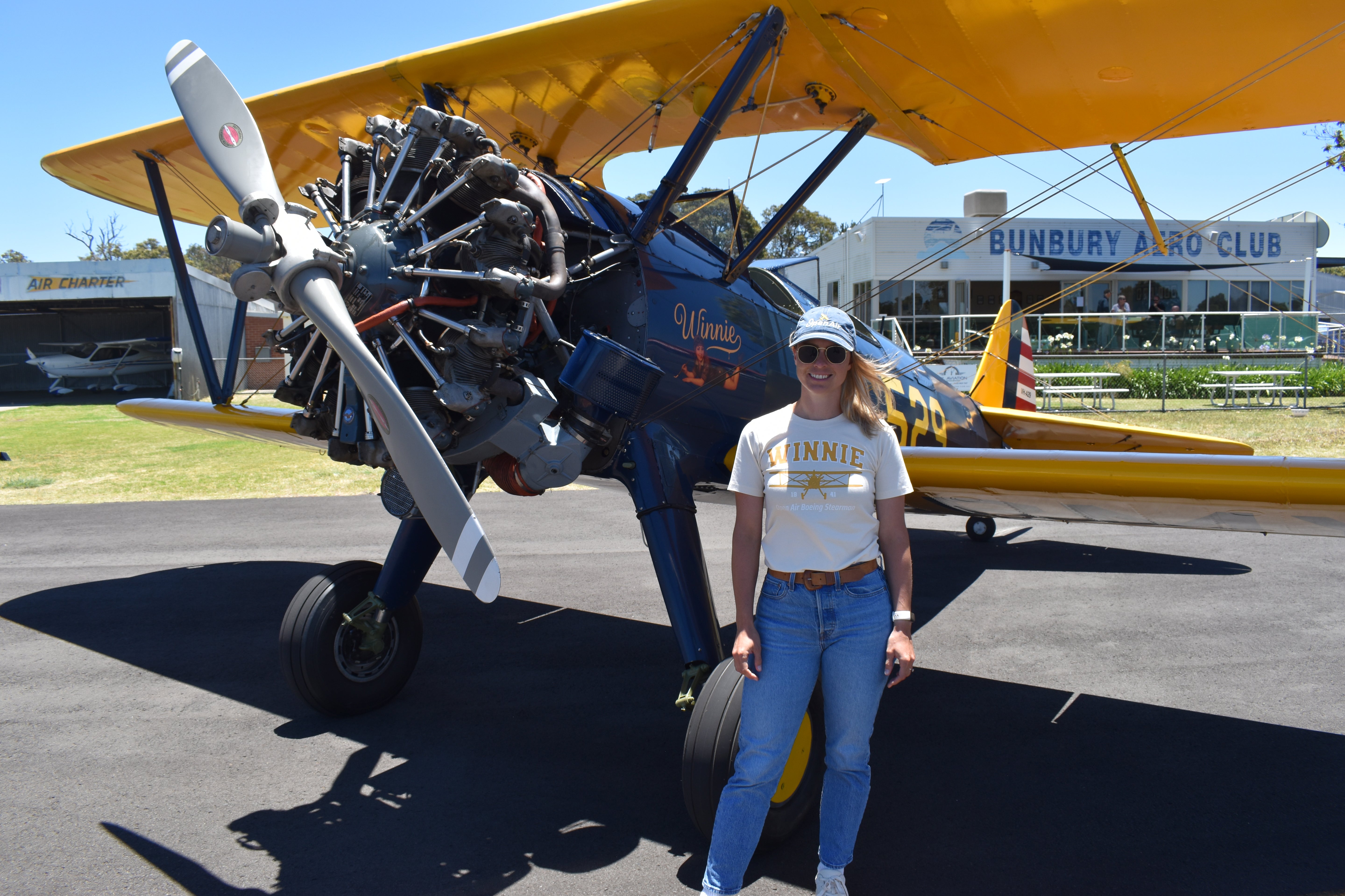 Hayley Coulson standing in front of Winnie, the restored 1941 Boeing Stearman, outside the Bunbury Aero Club wearing Open Air Adventure Flights merchandise.