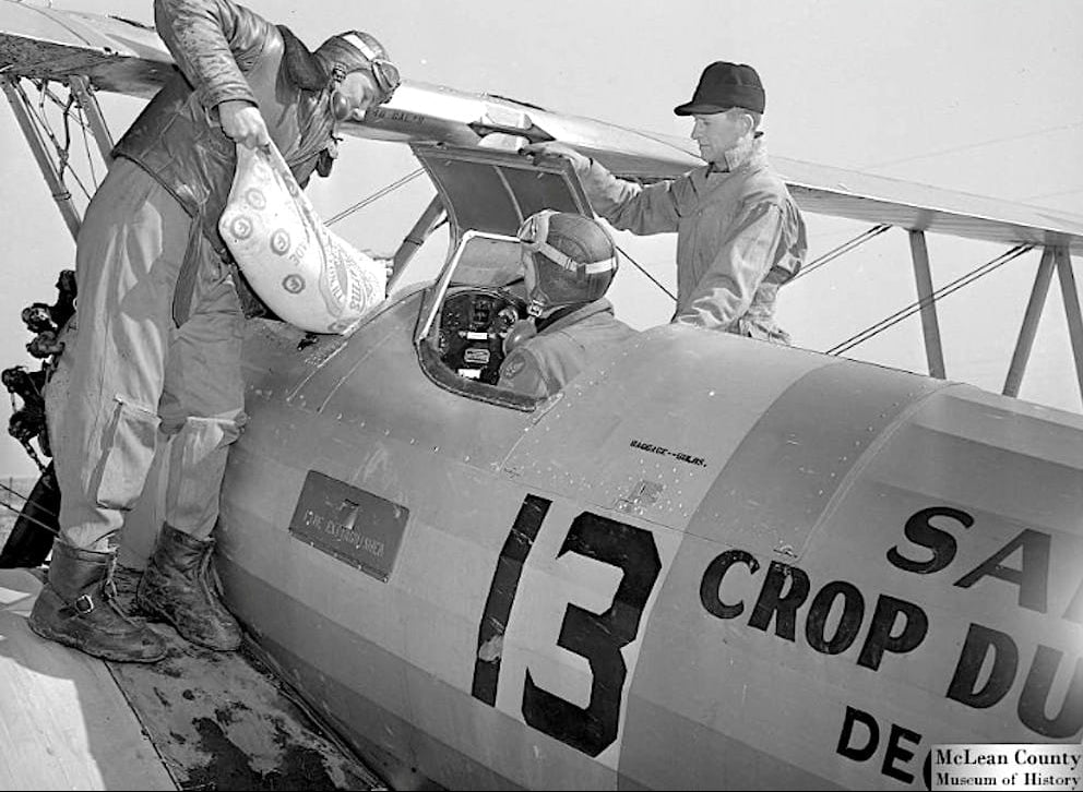 A vintage black and white photograph showing a Boeing-Stearman crop duster biplane marked with the number 13 being loaded for operation. One ground crew member stands on the lower wing pouring a large sack of chemical powder or seed into the hopper, while the pilot in a flight helmet watches from the open cockpit and another crew member assists from the side.