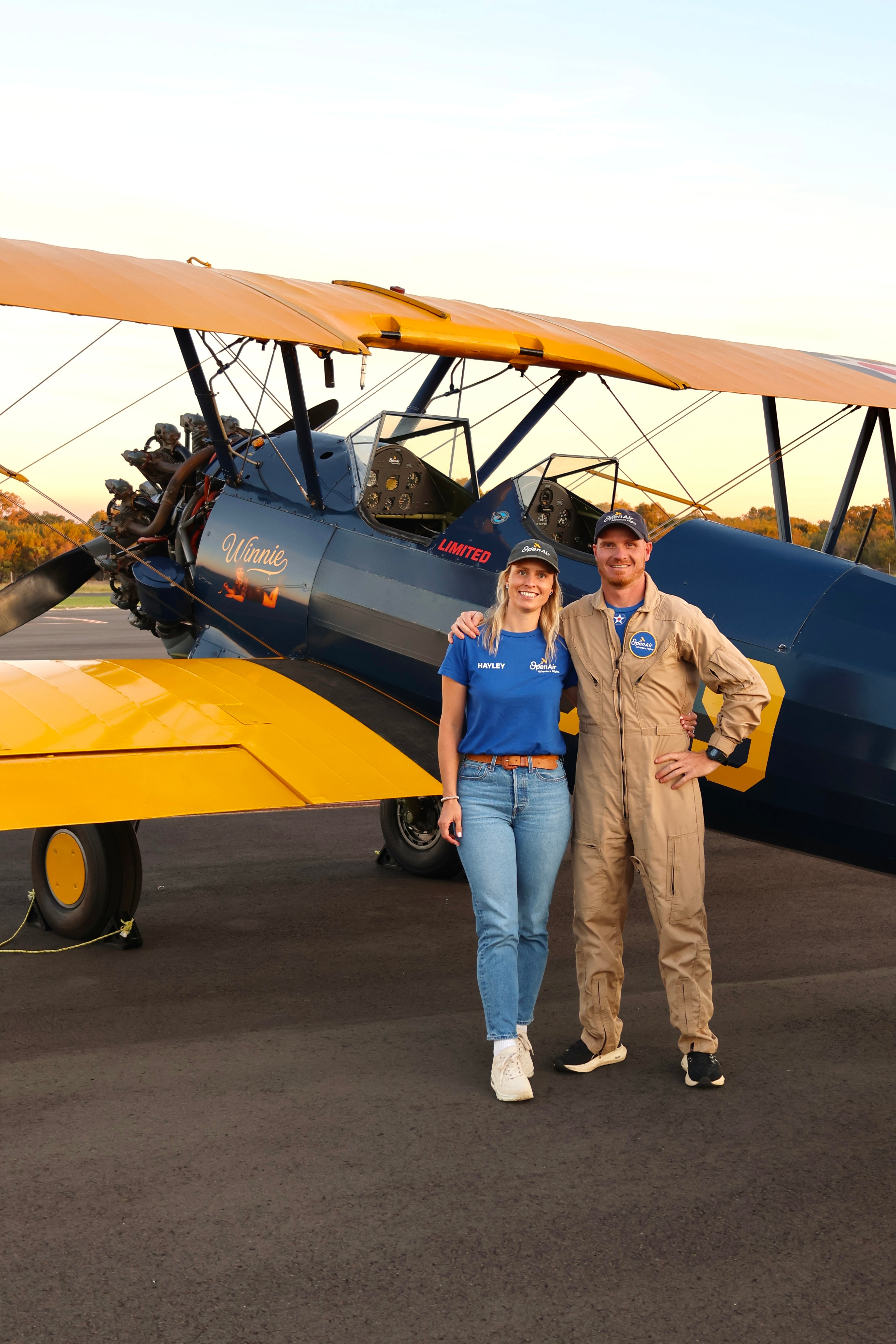 Pilot in flight suit standing with woman on airport tarmac next to yellow and blue Boeing-Stearman biplane.