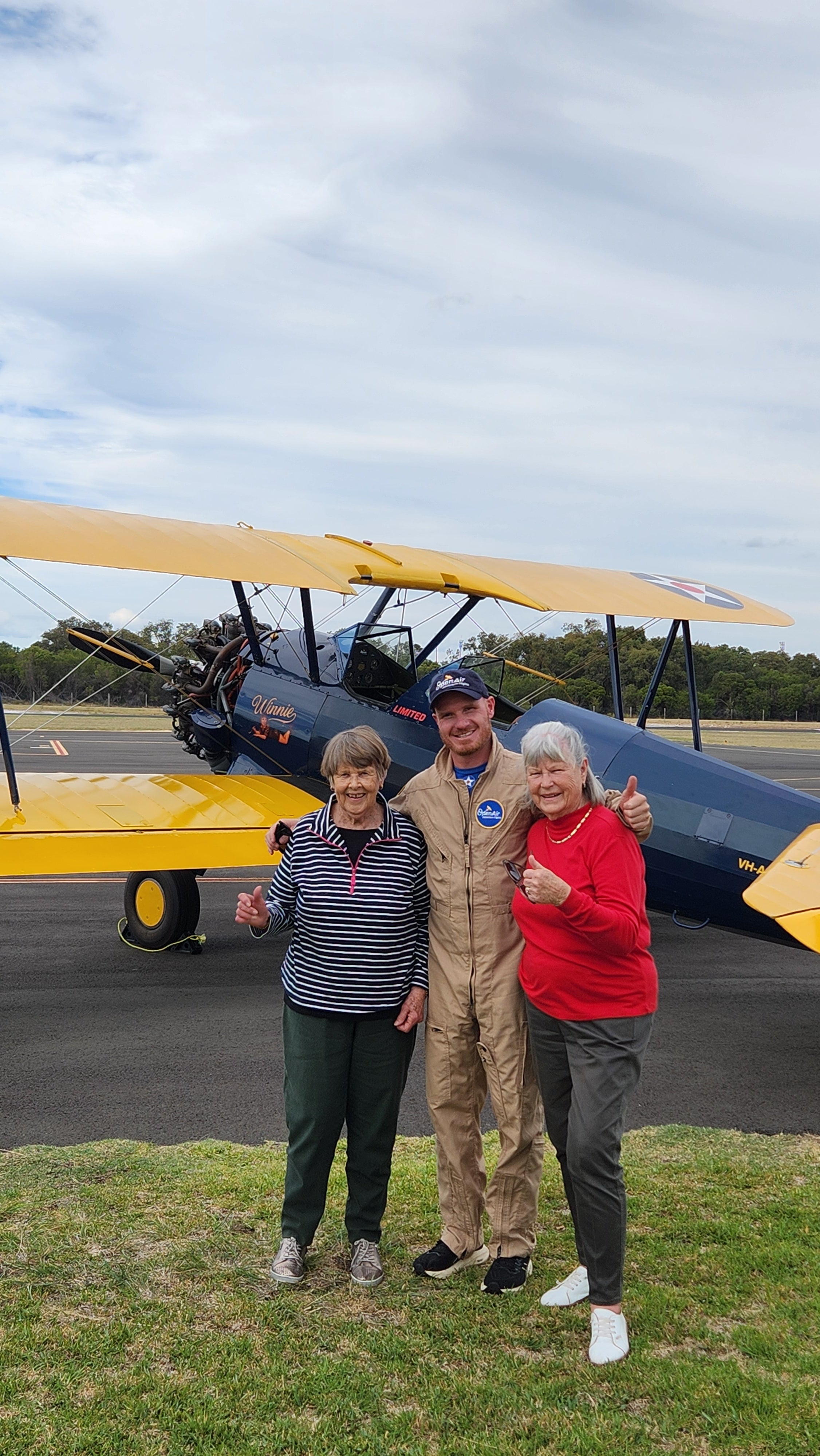 Pilot in a flight suit standing with two passengers next to a yellow and blue Boeing-Stearman biplane.