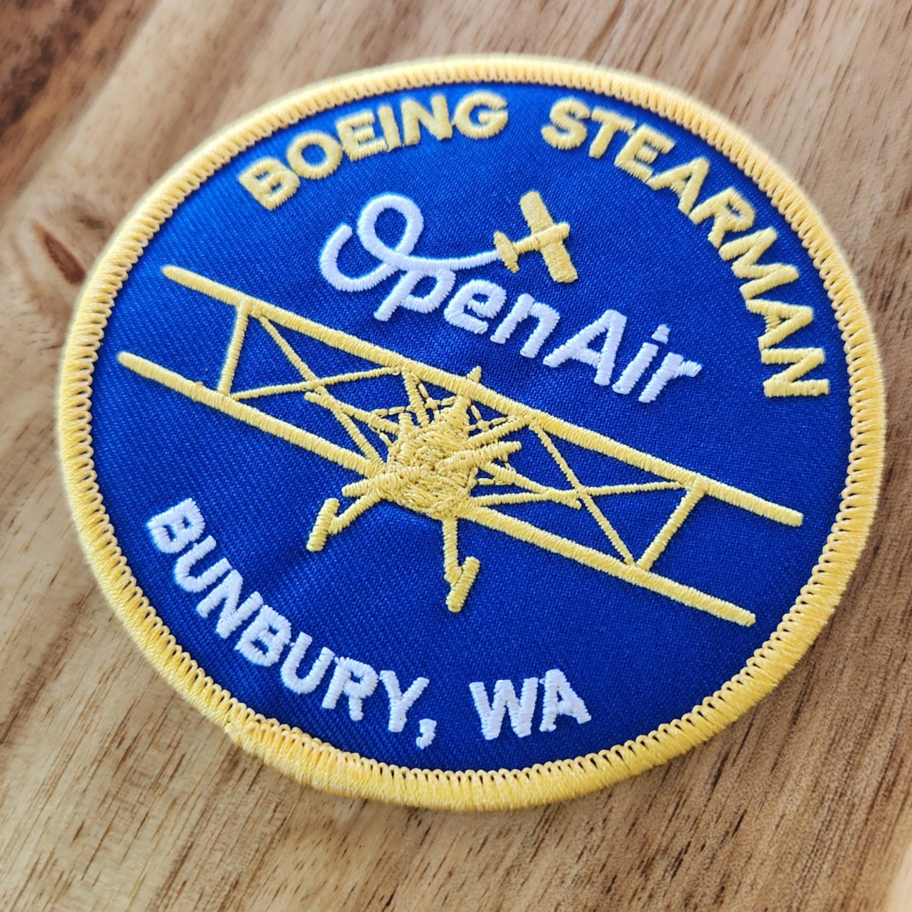 Open Air Adventure Flights Bunbury souvenir iron-on patch featuring a front-view Boeing Stearman, photographed on a wooden table.